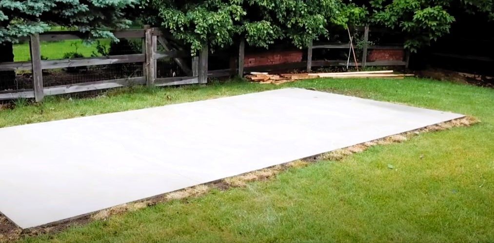 Freshly poured concrete slab in a grassy backyard with a wooden fence and trees