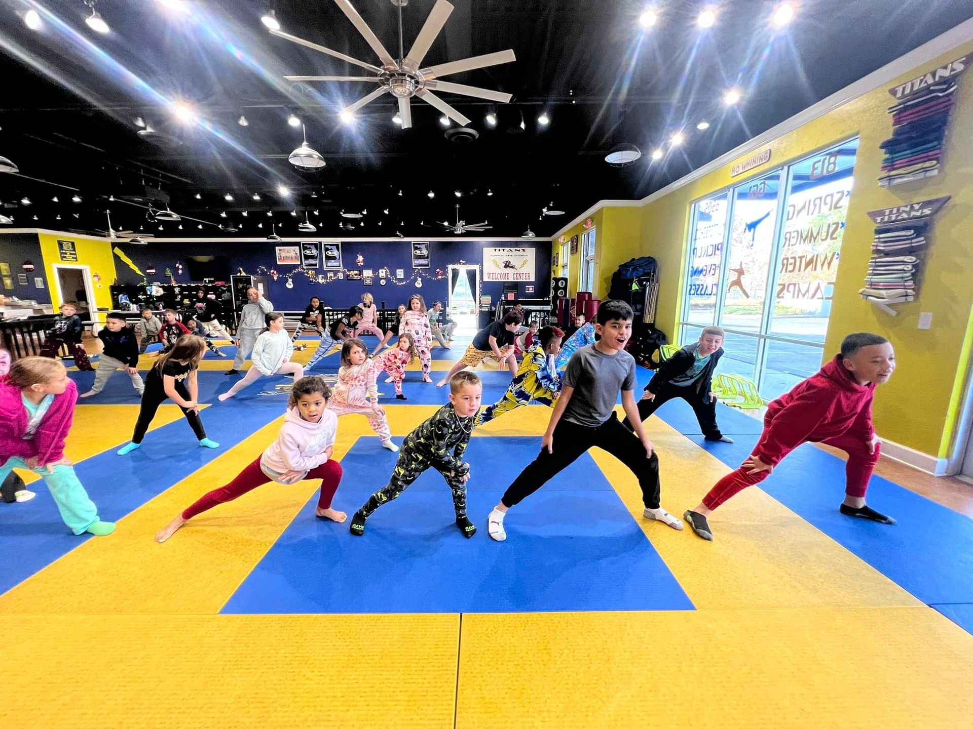 A group of children are doing stretching exercises on a blue and yellow mat in a gym.