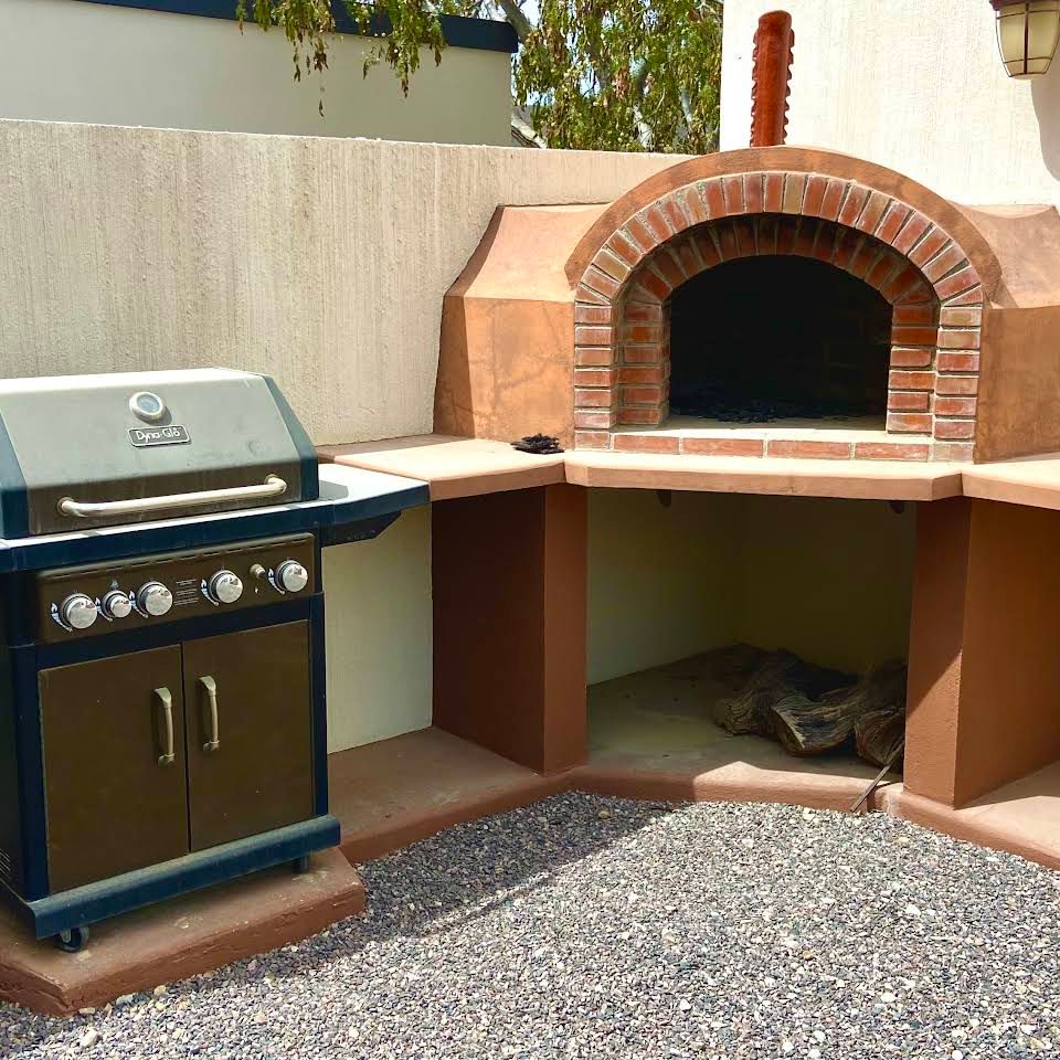 Outdoor patio with a gas grill beside a brick wood-fired pizza oven over a gravel ground