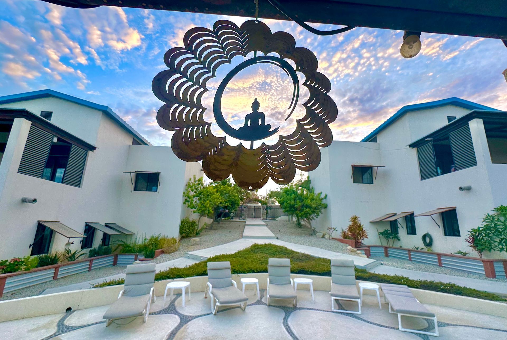Courtyard between white villas with lounge chairs, path, and ornate hanging ring sculpture at sunset.