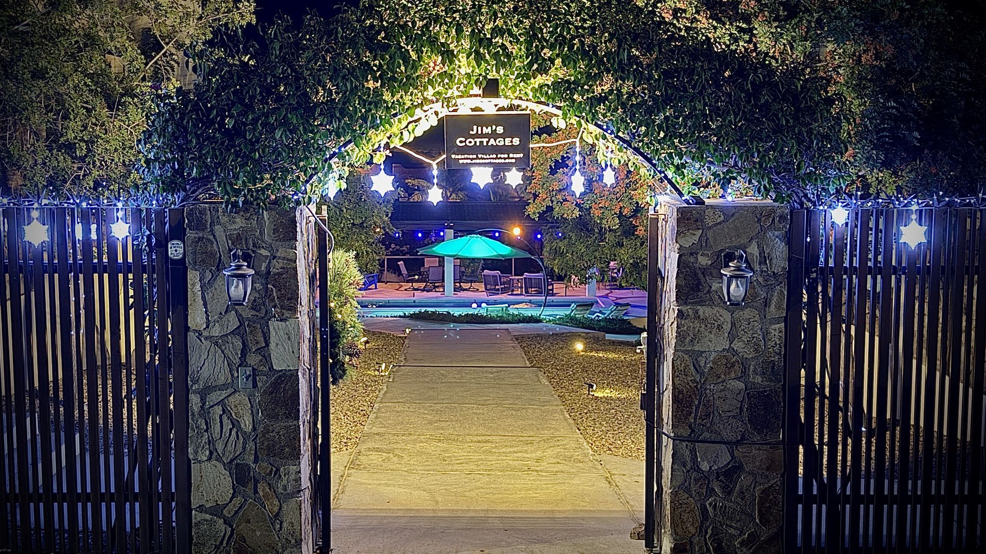 Lit garden entrance with stone pillars, open gates, and glowing lights leading to a decorated outdoor venue.