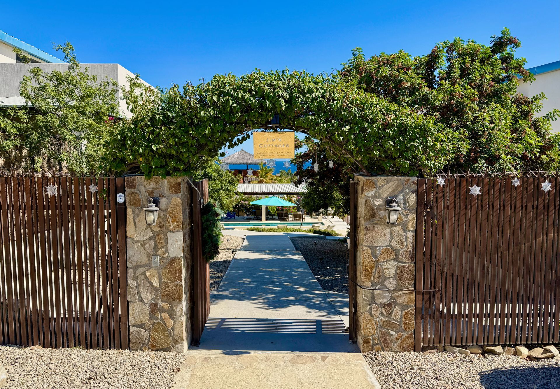 Stone gate with wooden slats opening to a sunny path under green vines, leading to a pool area
