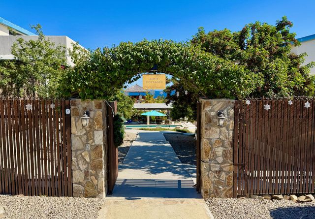Stone gate with wooden slats opening to a sunny path under green vines, leading to a pool area