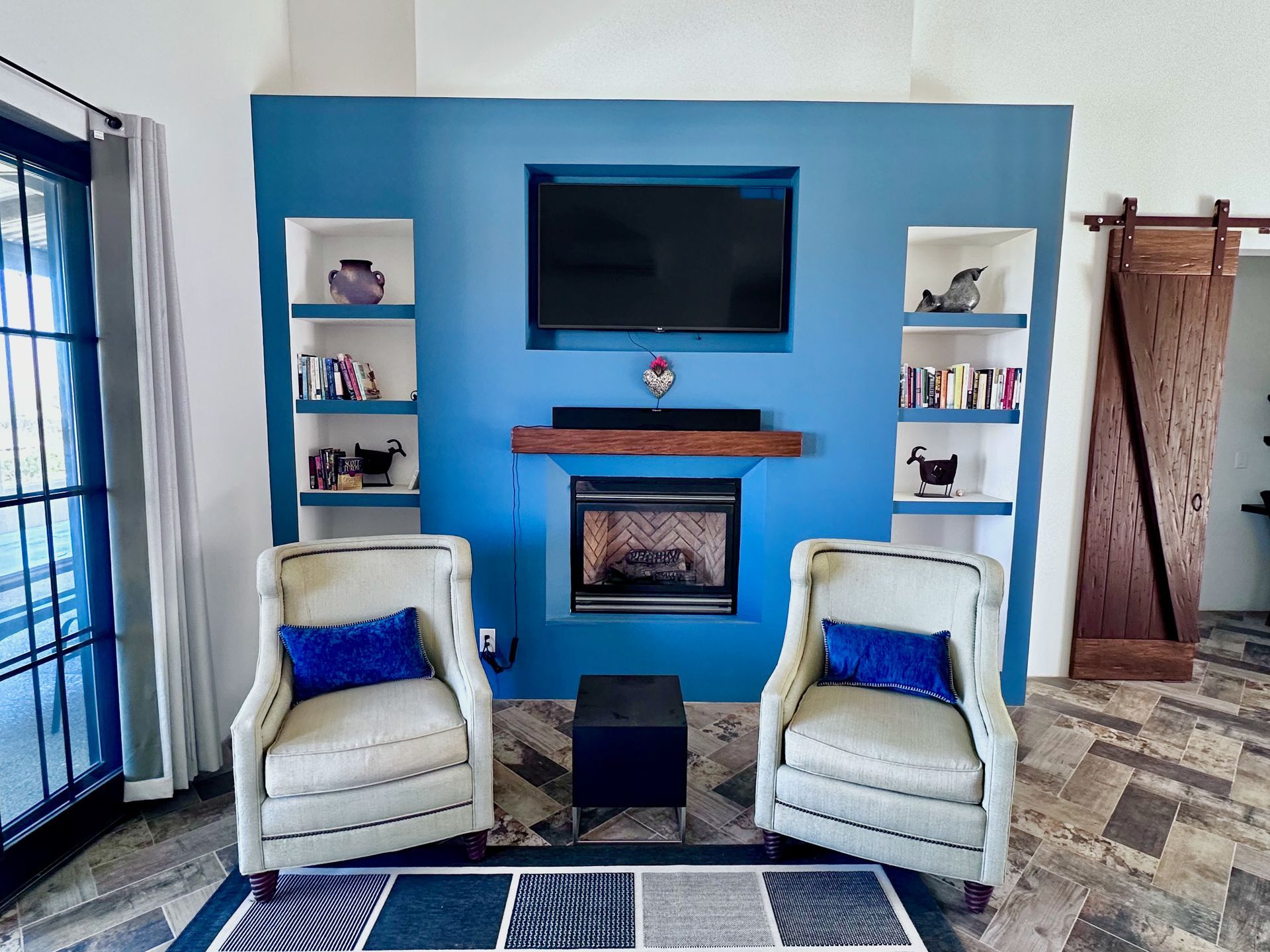 Blue living room with wall-mounted TV, fireplace, two white chairs, and sliding glass doors