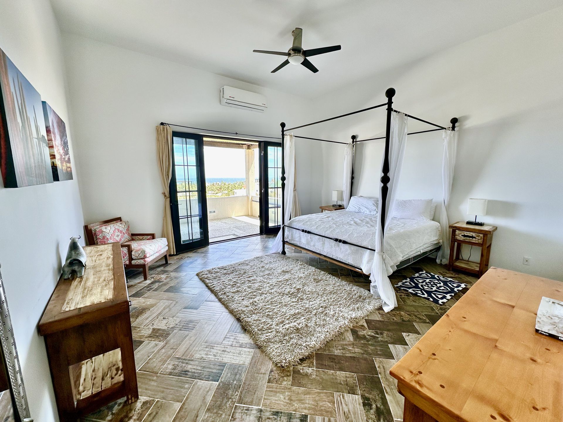 Bright bedroom with a canopy bed, wooden furniture, patterned rug, and open doors to a balcony.