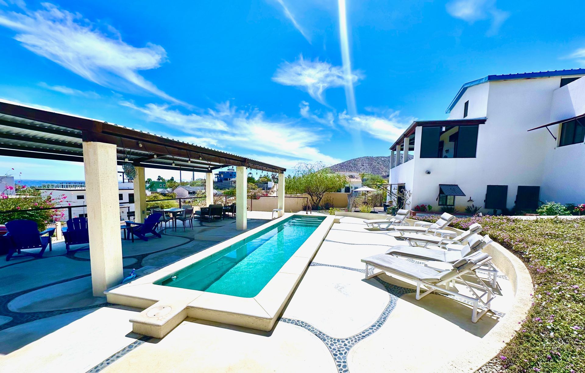 Modern patio with a lap pool, white lounge chairs, and a white house under a bright blue sky