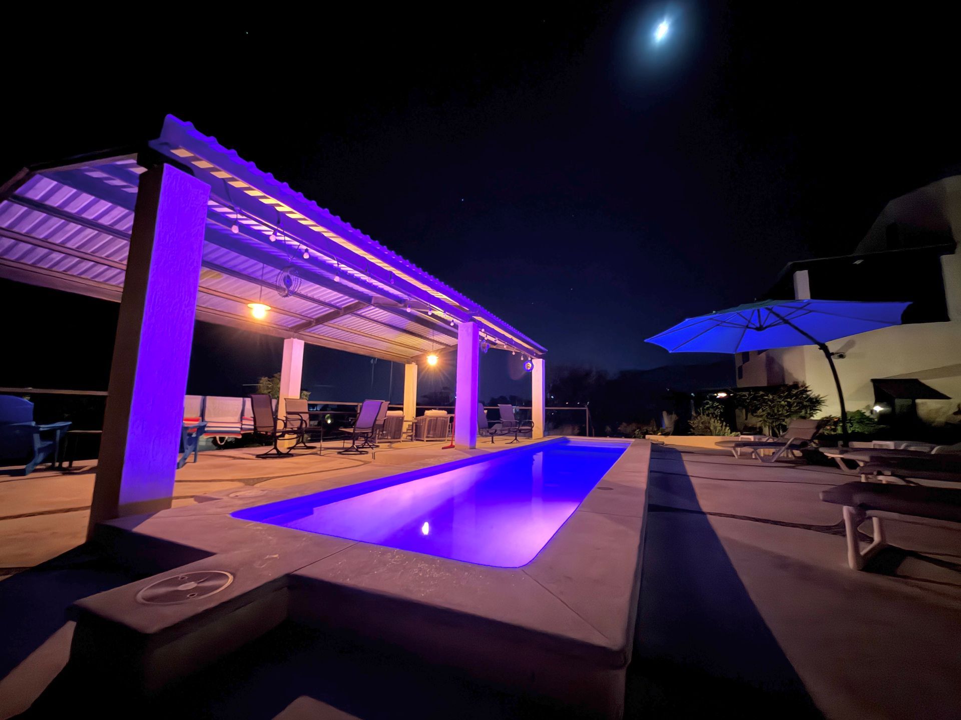 Purple-lit backyard pool and patio at night under a pergola, with lounge chairs and moon overhead