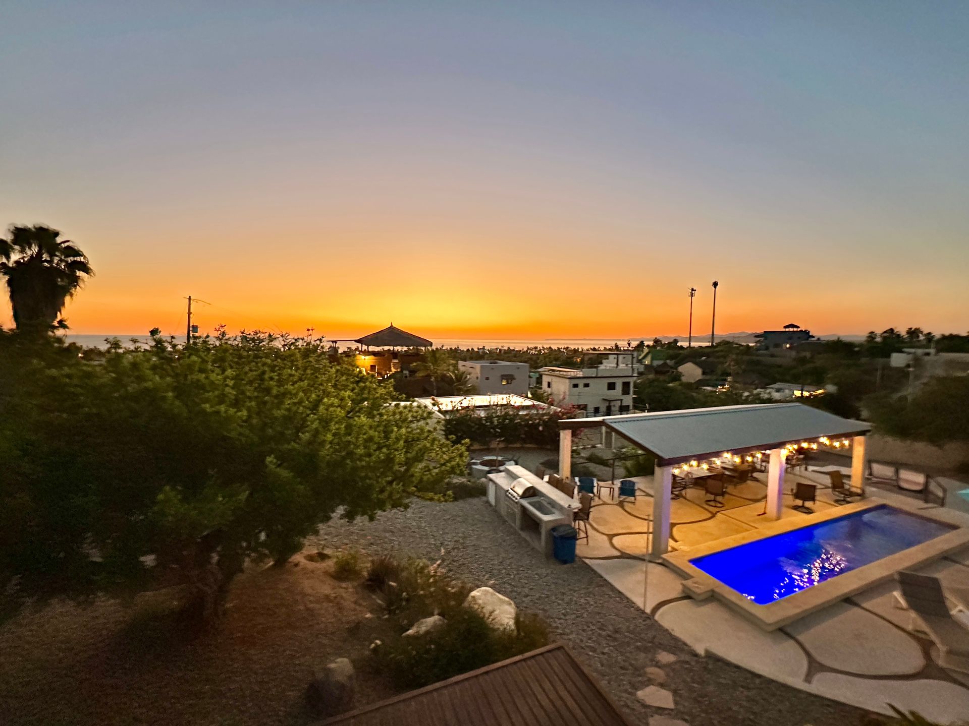 Sunset over a villa with a lit blue pool, patio, and palm trees overlooking rooftops