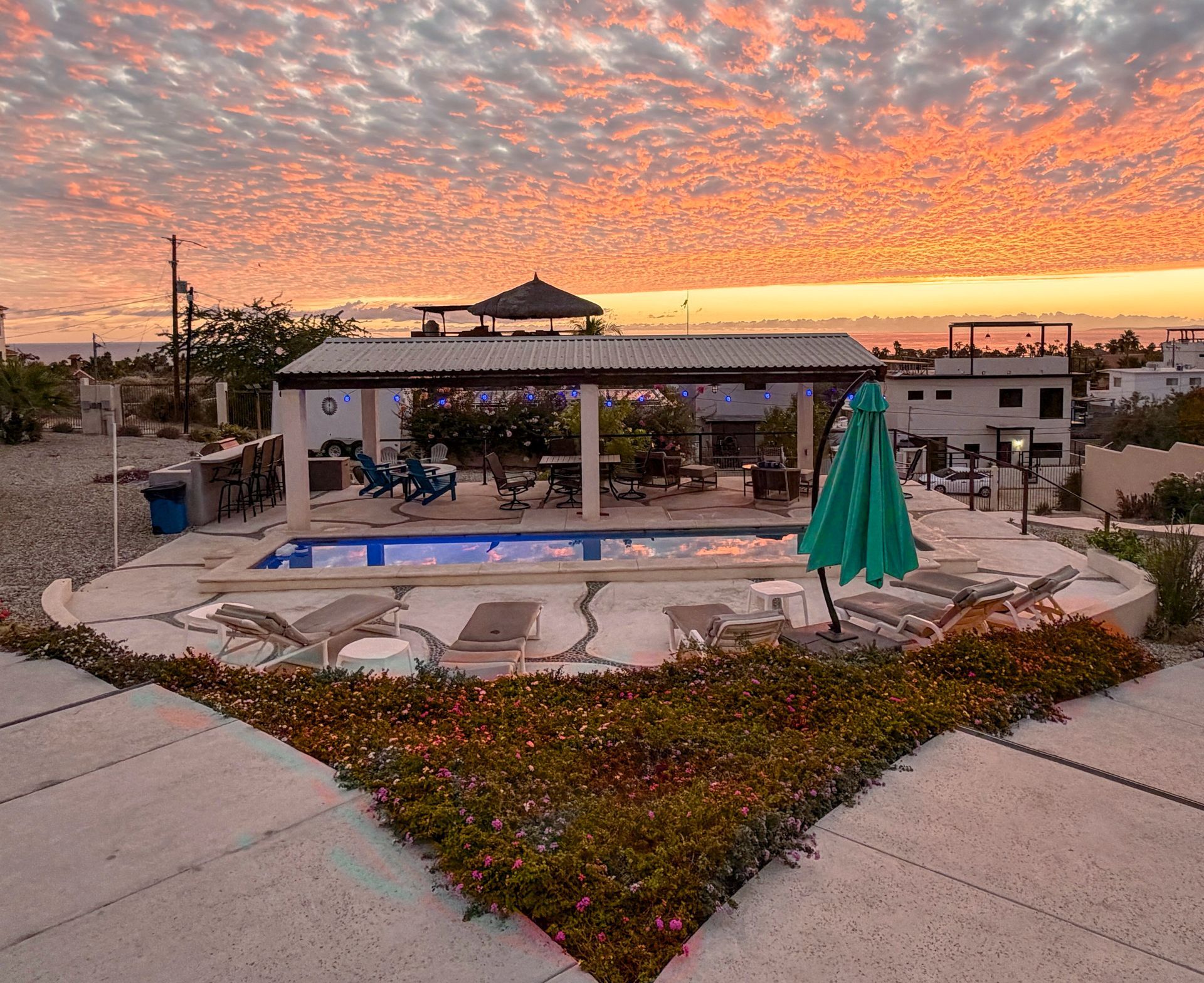 Poolside patio with lounge chairs and a green umbrella at sunset under a pink-orange sky