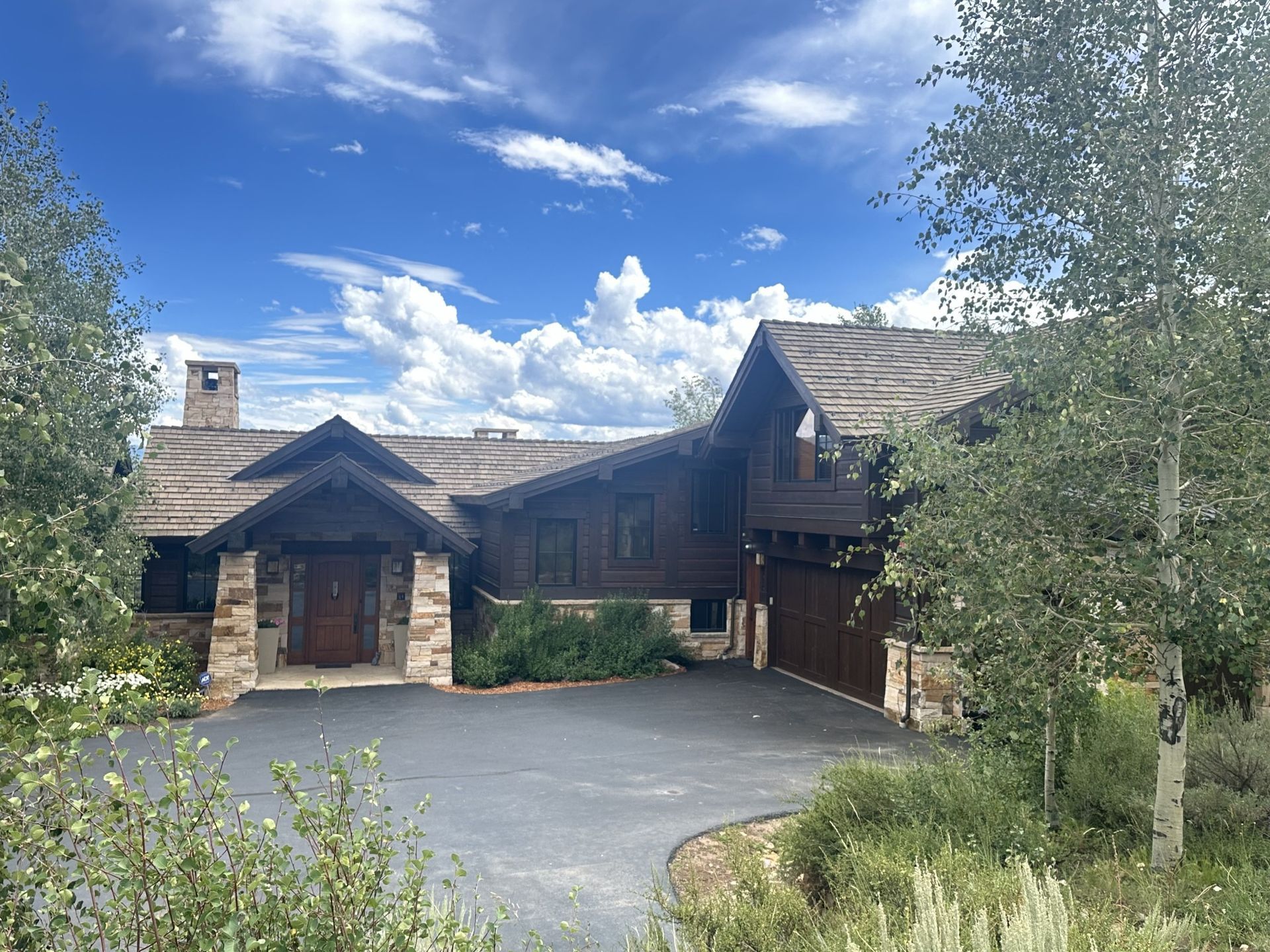 Large rustic house with stone and wood exterior, driveway, and trees under a partly cloudy blue sky