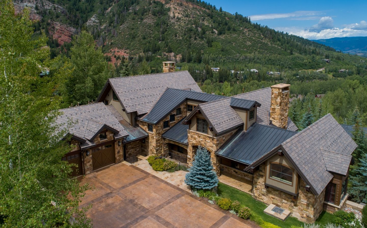 Large rustic house with stone and wood exterior, driveway, and trees under a partly cloudy blue sky