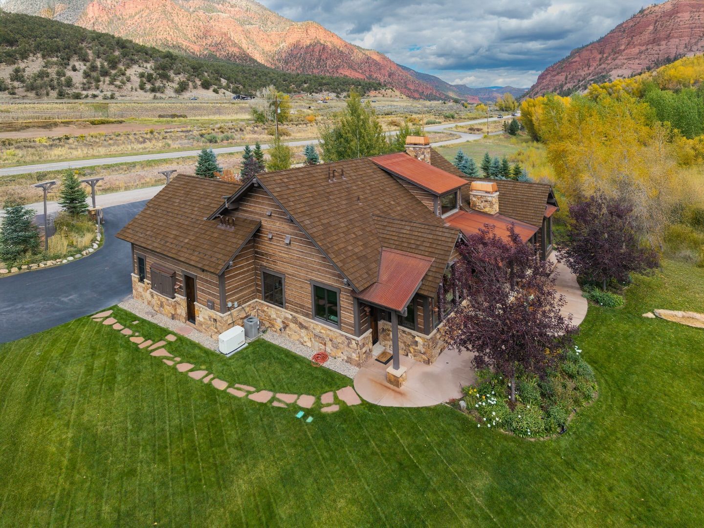 Rustic house with brown roof by a river, green lawn, and autumn trees with mountains in the background