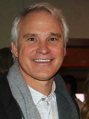 Smiling man in a gray jacket and patterned white shirt, posing indoors.