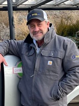Man in a gray jacket and cap leaning on a boat deck, standing beside marsh grass and water.