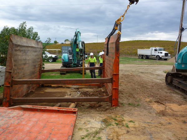 Construction workers near trench box at a construction site, with excavator, truck, and cloudy sky.
