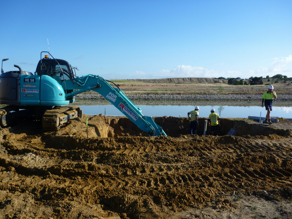 An excavator digs a trench next to a body of water, with workers nearby under a clear blue sky.