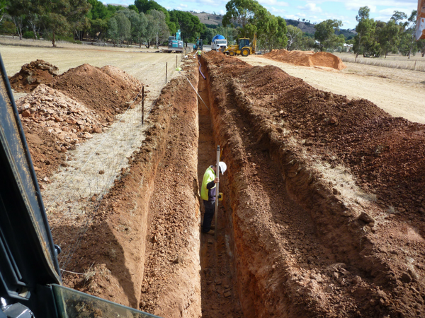 Worker in safety vest and hard hat inspecting a long, narrow trench at a construction site.