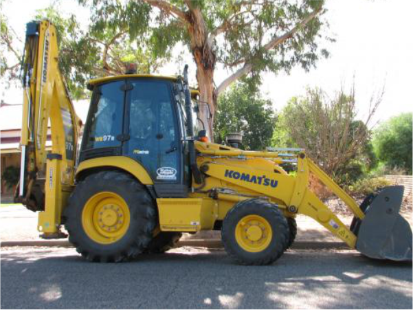 Yellow Komatsu backhoe loader parked on a paved road near a tree.