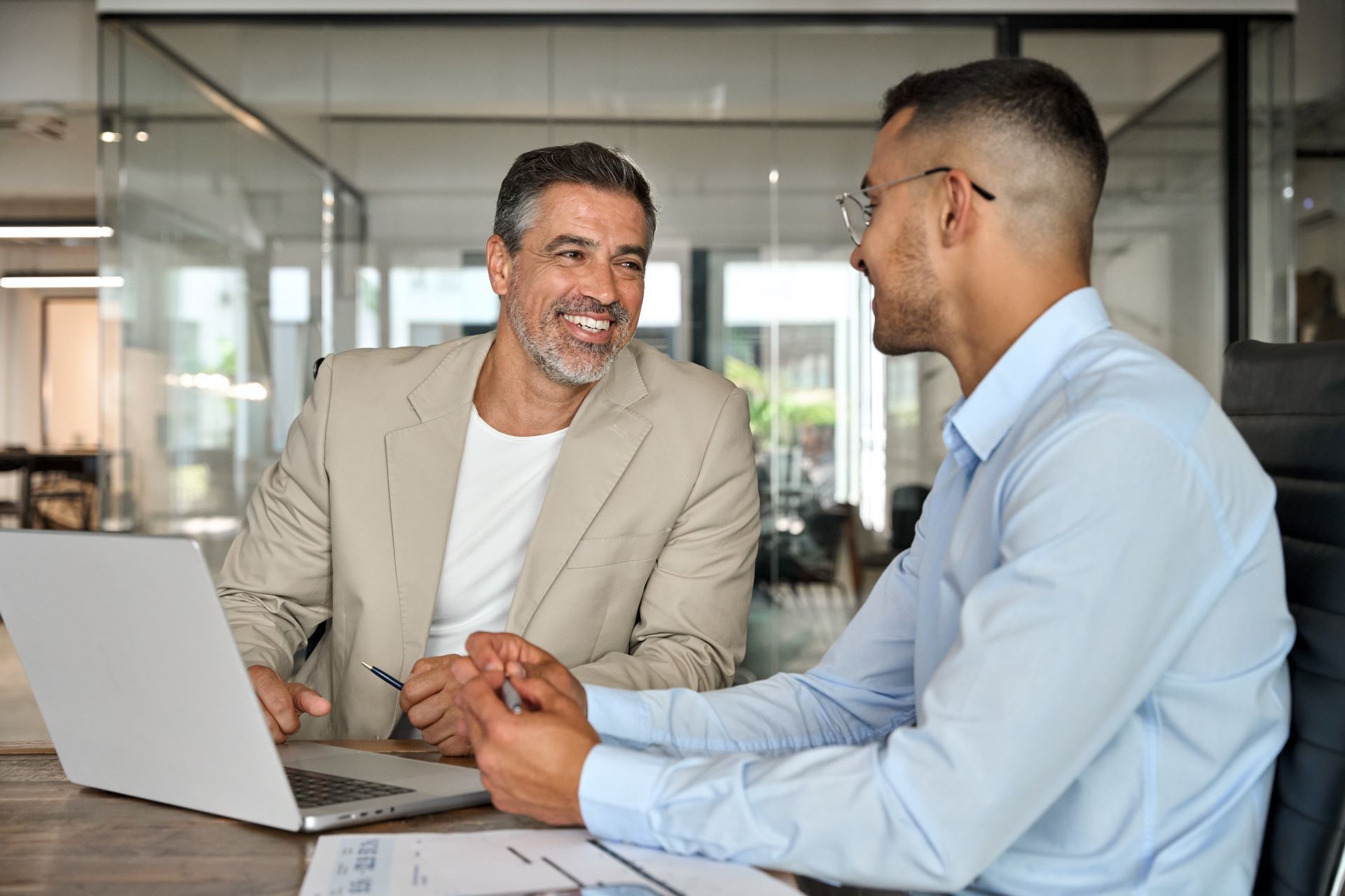 Two colleagues smiling and talking at a conference table with a laptop in a glass-walled office