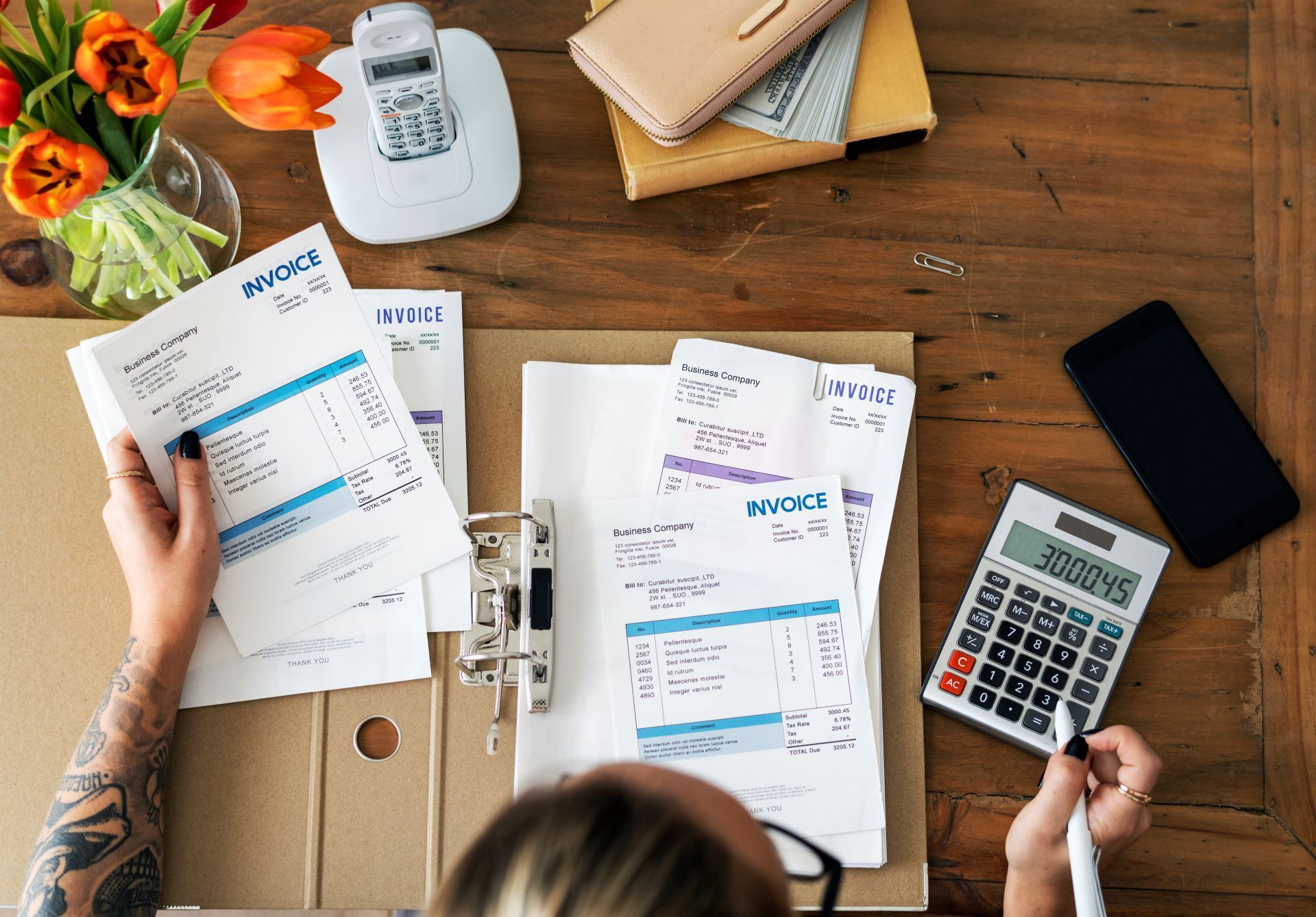 Person reviewing invoices, surrounded by bills, calculator, phone, and flowers on a wooden desk.