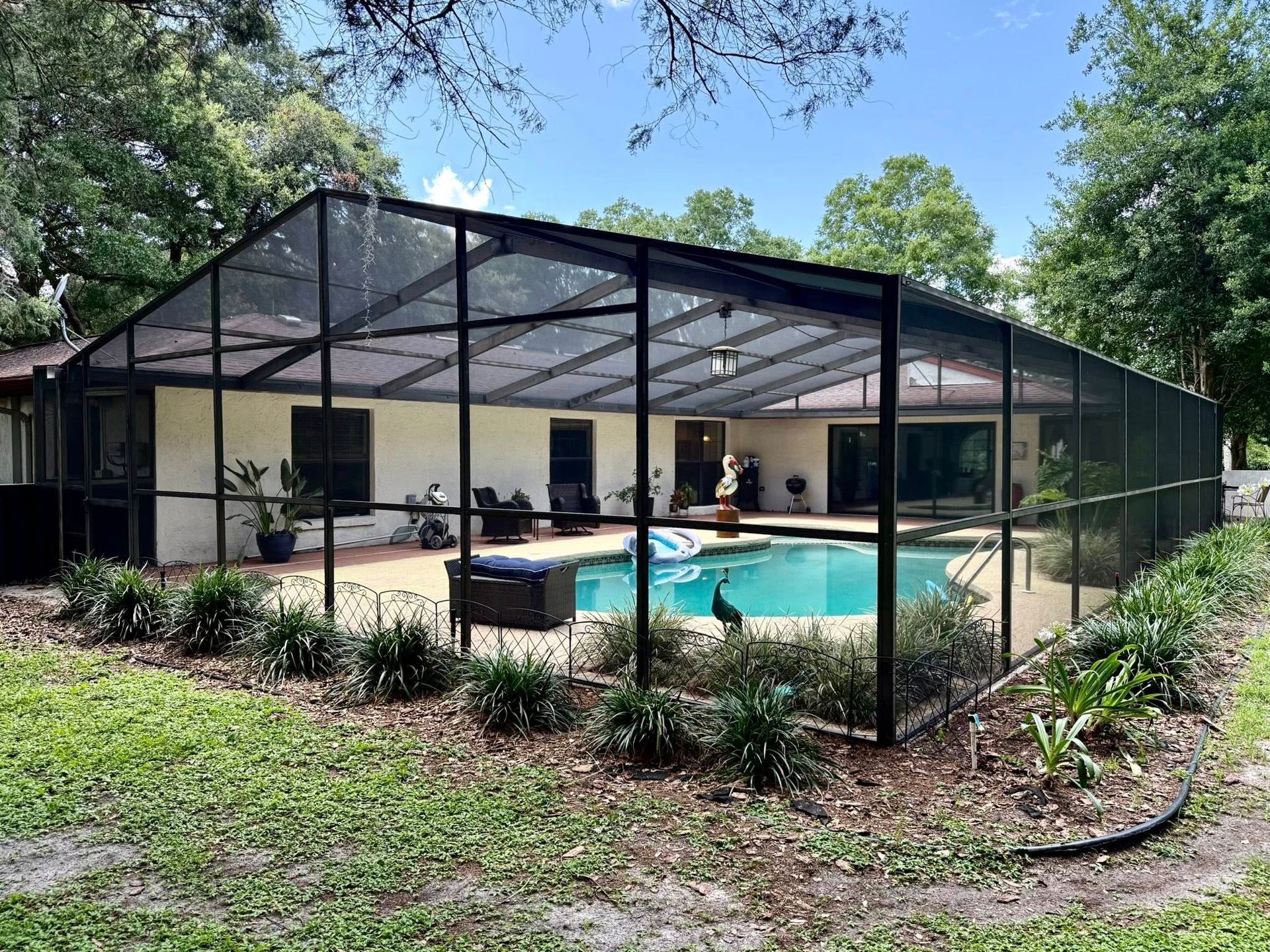 Pool enclosed in black mesh with beige house. Lush greenery in yard, sunny day.