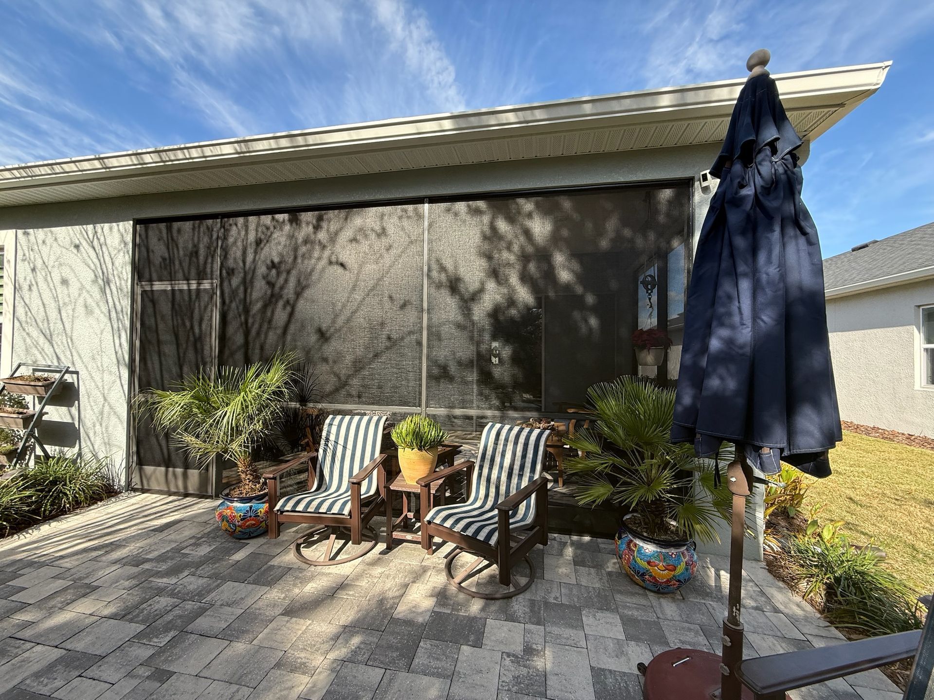 Patio with two striped chairs, potted plants, and a closed navy umbrella beside a house.