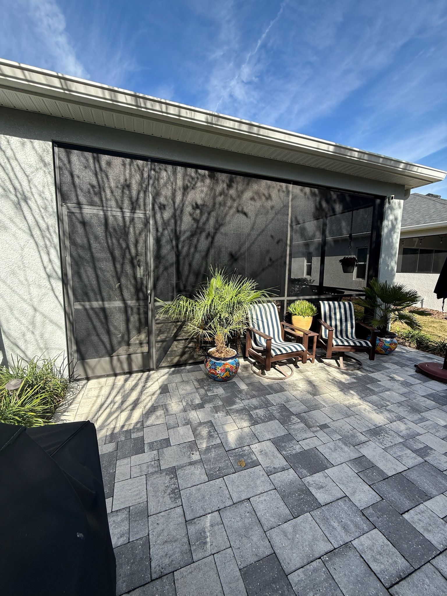 Patio with gray pavers, screened wall, and potted plants beside a house and outdoor chairs
