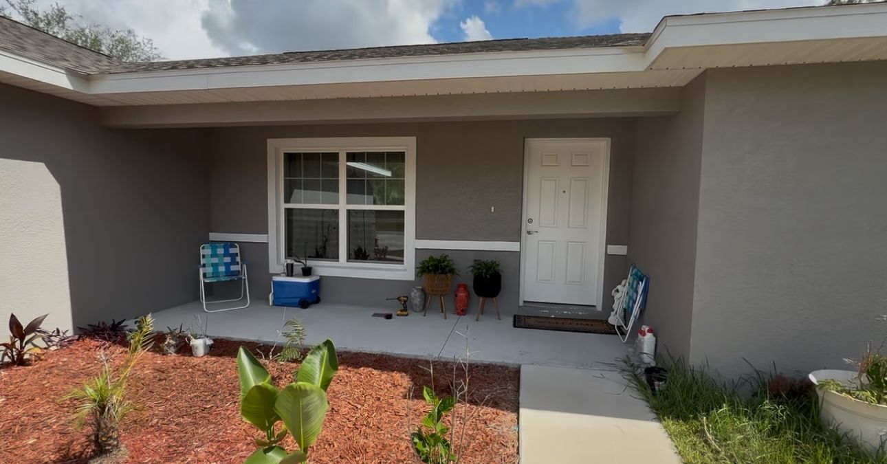 Gray house with white door and window, porch with chairs, plants, and blue cooler.