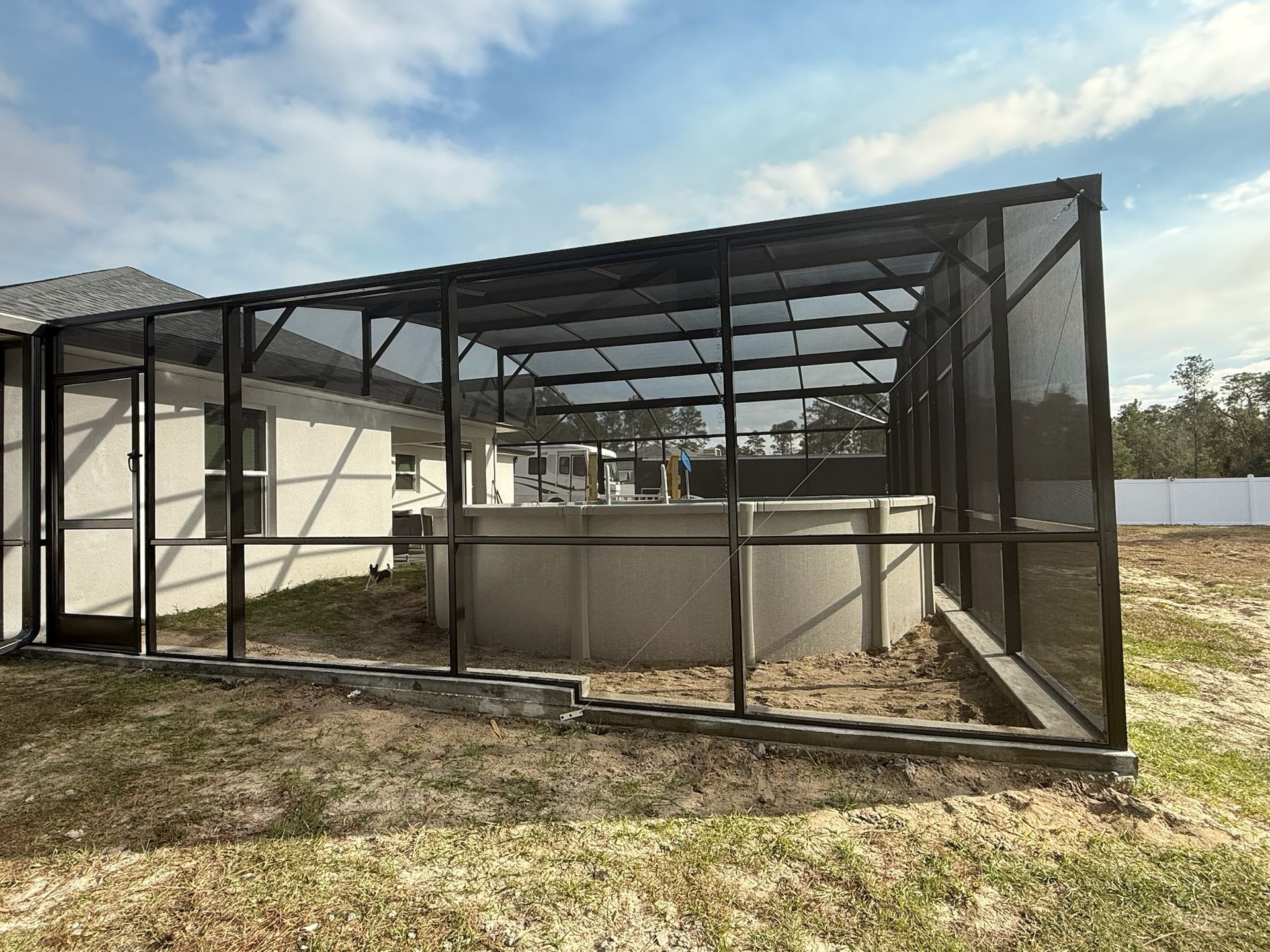 Black mesh-enclosed patio or pool cage beside a house in a dry yard under a clear sky
