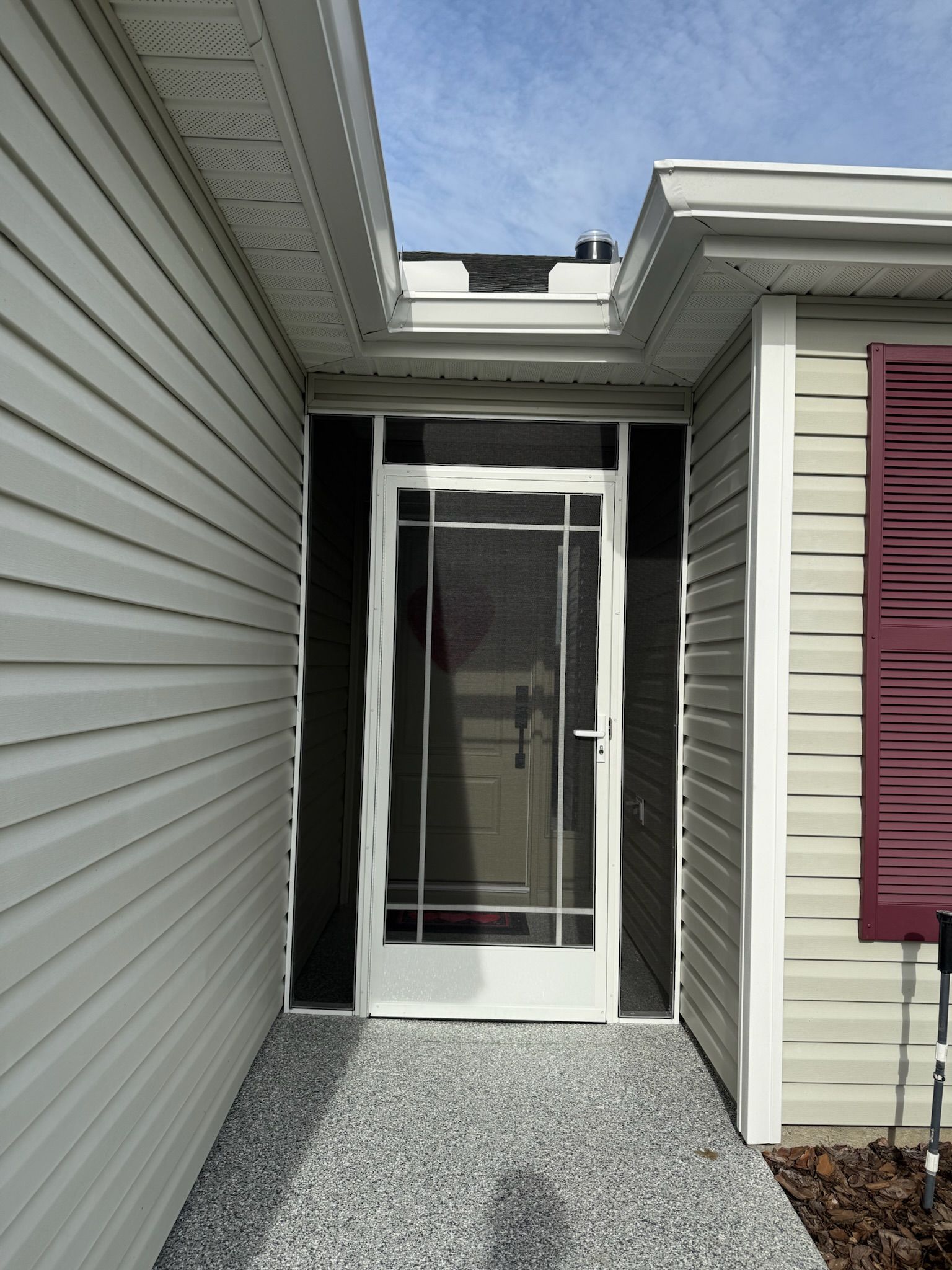 Front entry with a white screen door between gray siding and beige walls, with a maroon shutter on the right.