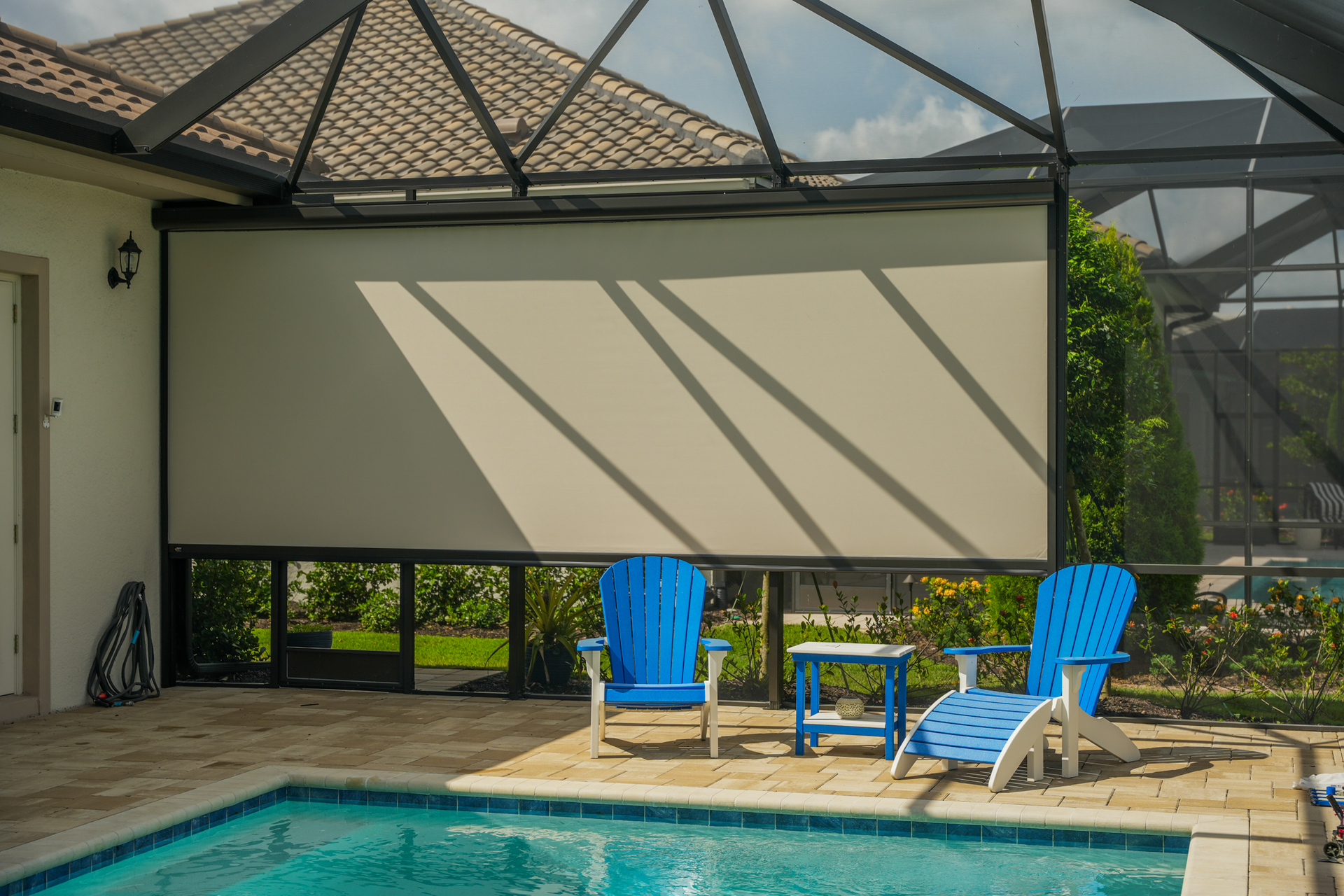 Screened patio by a pool with beige shade screen and two blue chairs beside a small table