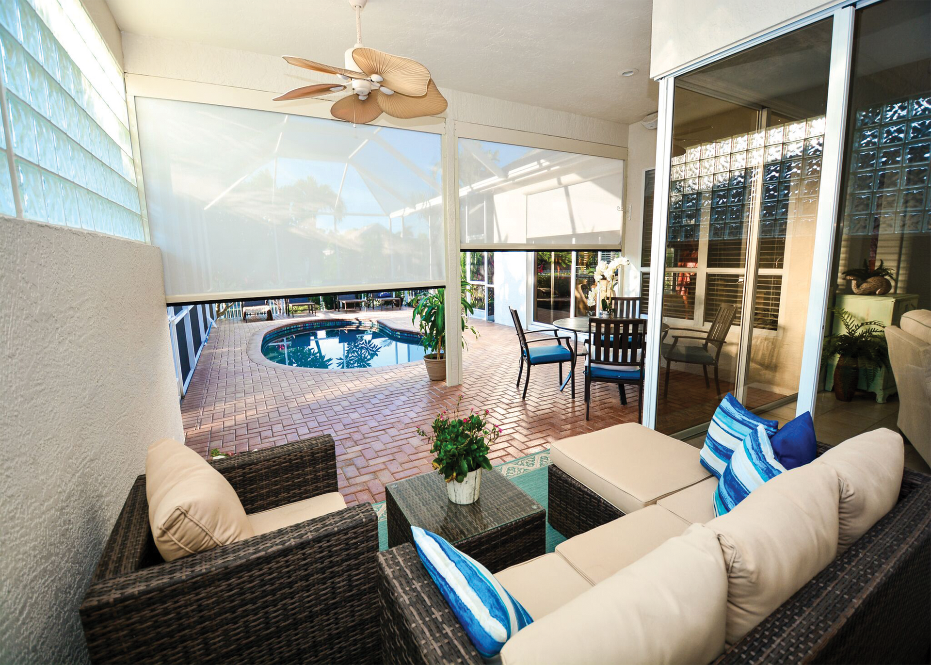 Covered patio lounge with wicker seating, blue cushions, and a view of a swimming pool and ocean.