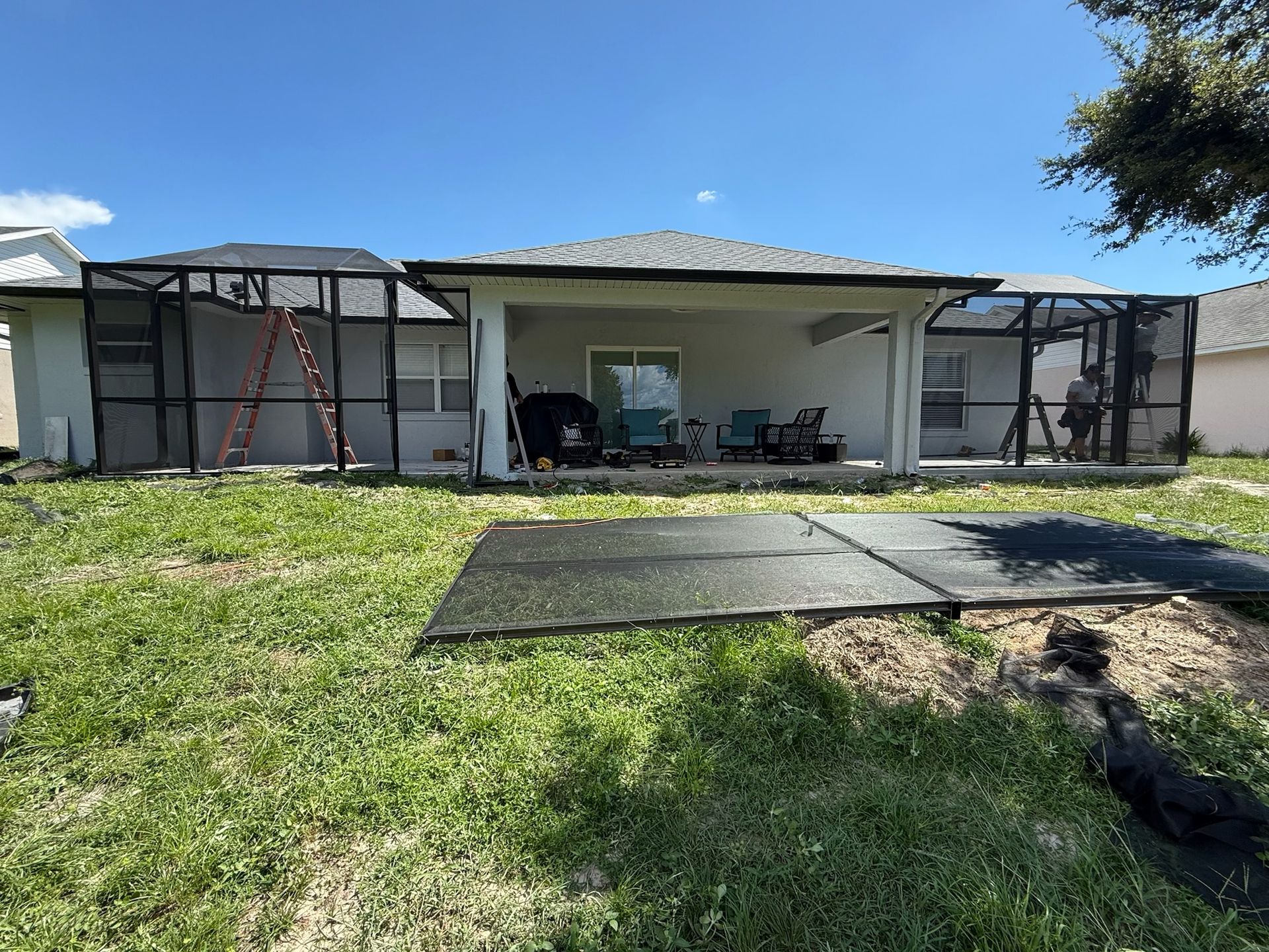 Backyard view of a house with a screened porch, ladder, and black mats on the lawn under a blue sky.