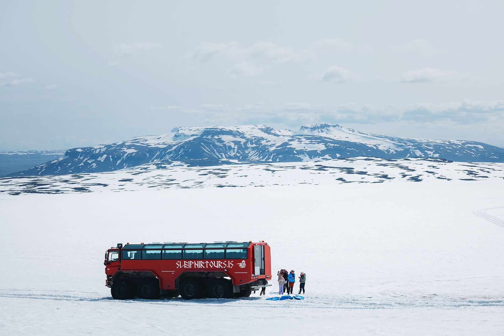 A giant red Sleipnir 8x8 monster truck parked on the white snow of Langjökull glacier under a clear blue sky.
