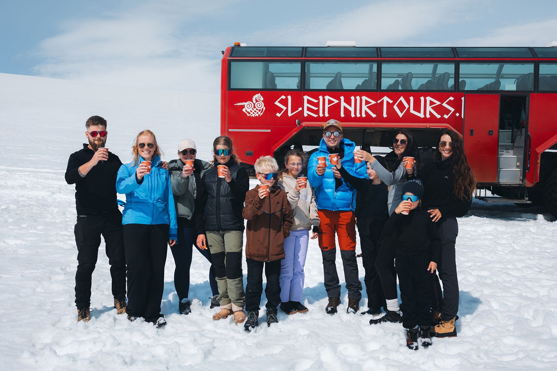 A group enjoying a cup of hot cocoa on Langjökull glacier, standing in front of the Sleipnir Monster truck.