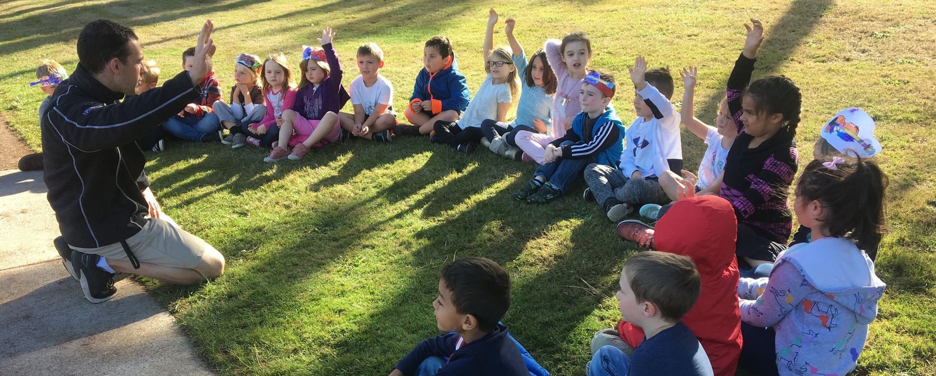 Diverse group of children with teacher on grass with hands raised to give answer.