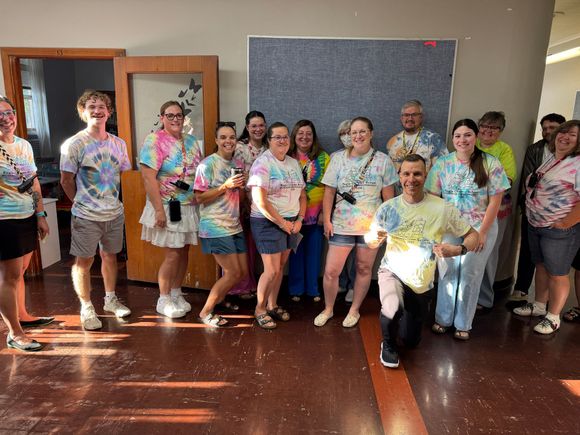 A group of 16 staff and teachers in a hall wearing tie-dye shirts made for summer enrichment.