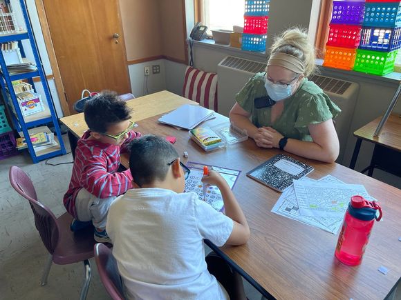 2 children with 1 teacher doing school work at a table in a classroom.