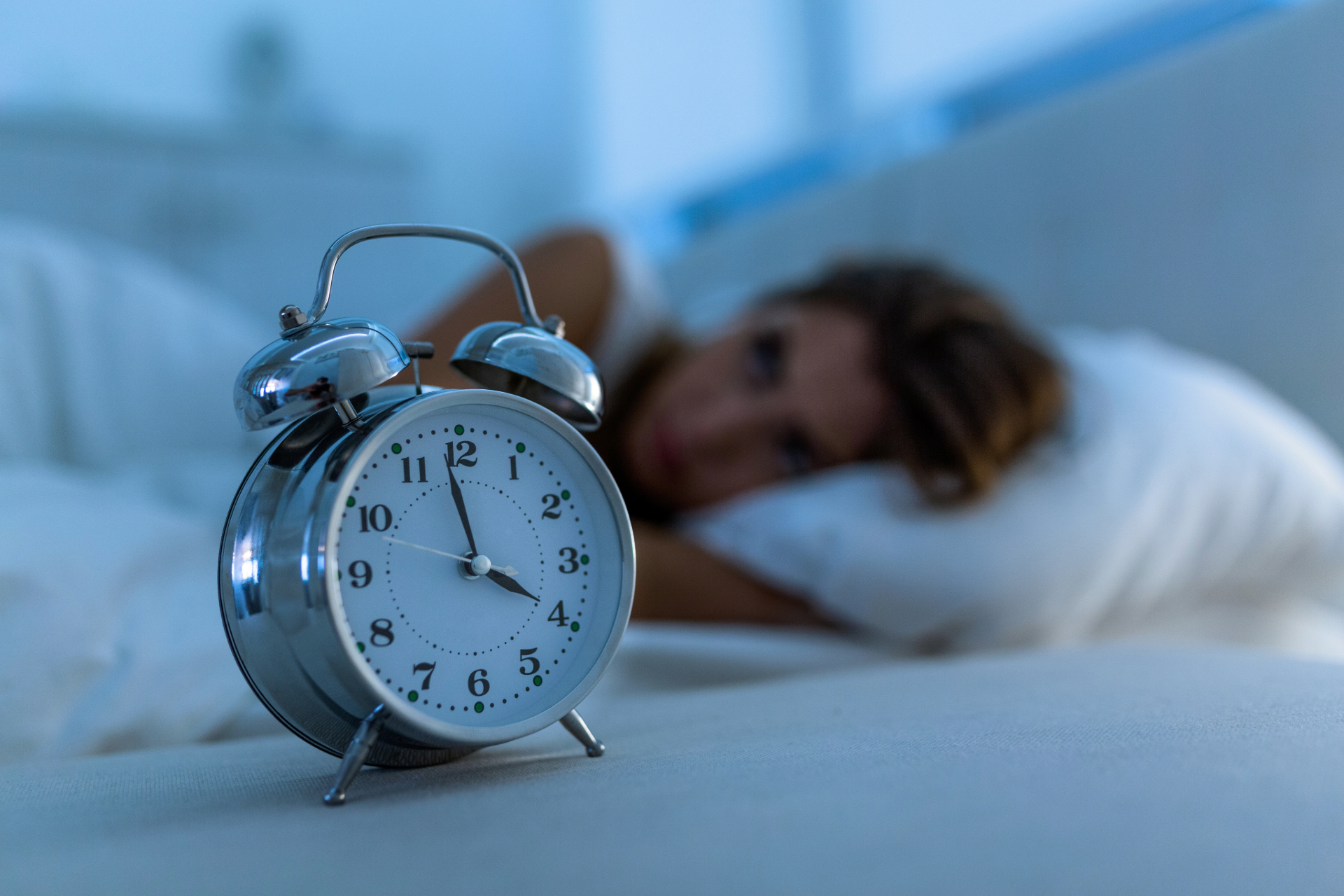 Alarm clock in focus on a bed, with a person sleeping blurred in the background.