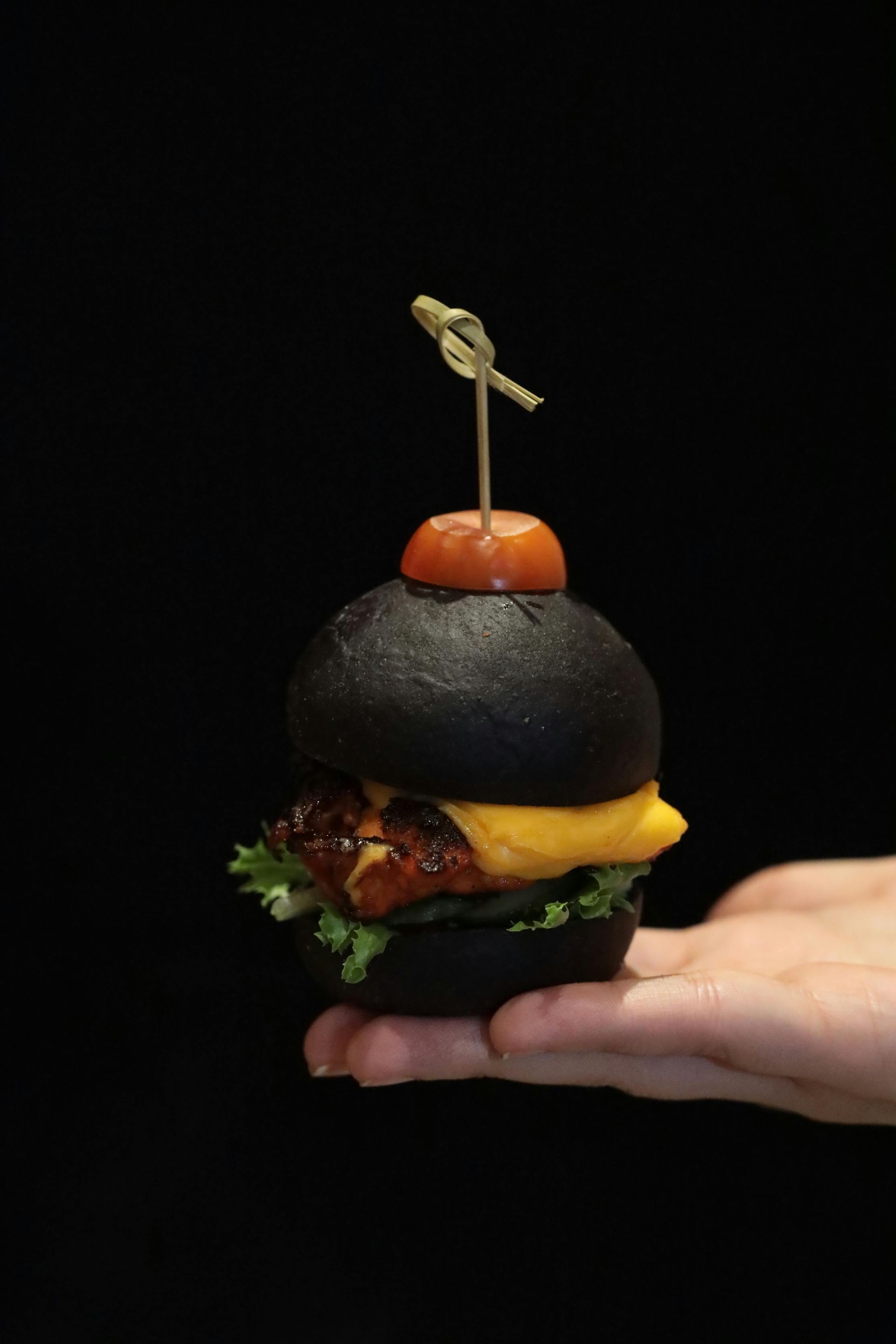 Hand holding a mini black burger topped with a cherry tomato and toothpick, against a black background.