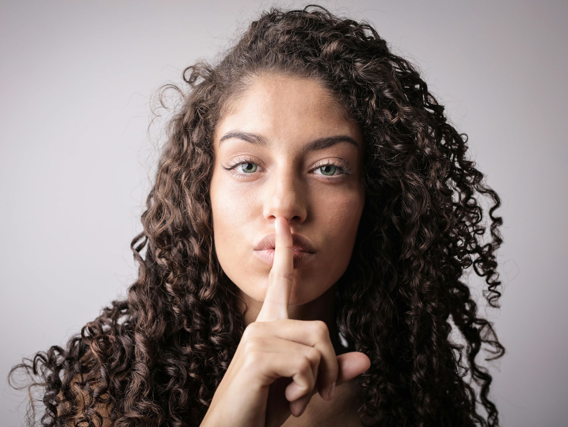 Woman with curly hair, finger to lips, gesturing for silence against a grey background.
