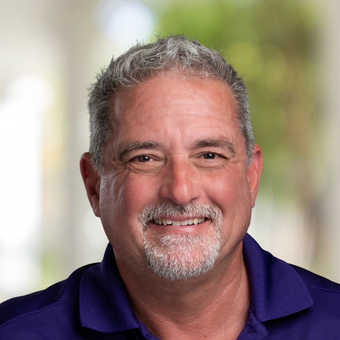 A man with a beard is wearing a purple shirt and smiling.