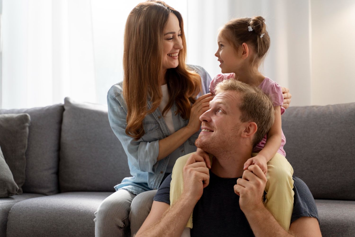 Smiling family of three sitting on a couch, with a child hugging their parents in a bright living room
