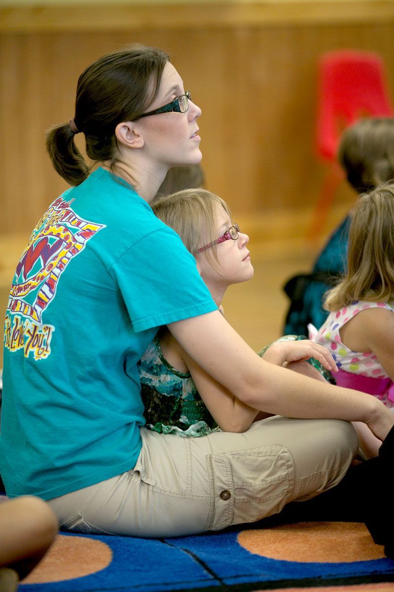 Woman sitting cross-legged on a carpet, holding a child, watching something off-screen in a classroom.
