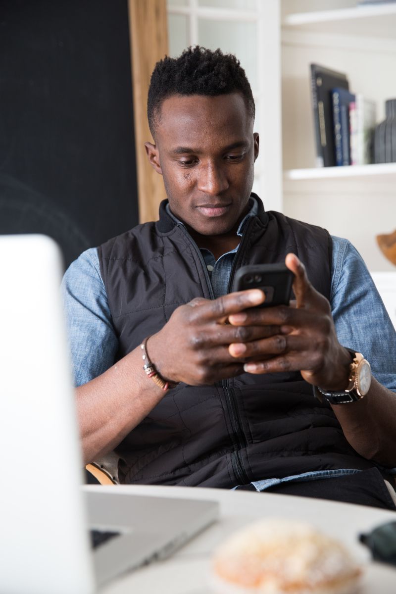 Man sitting on a couch, looking at a smartphone in a bright living room.