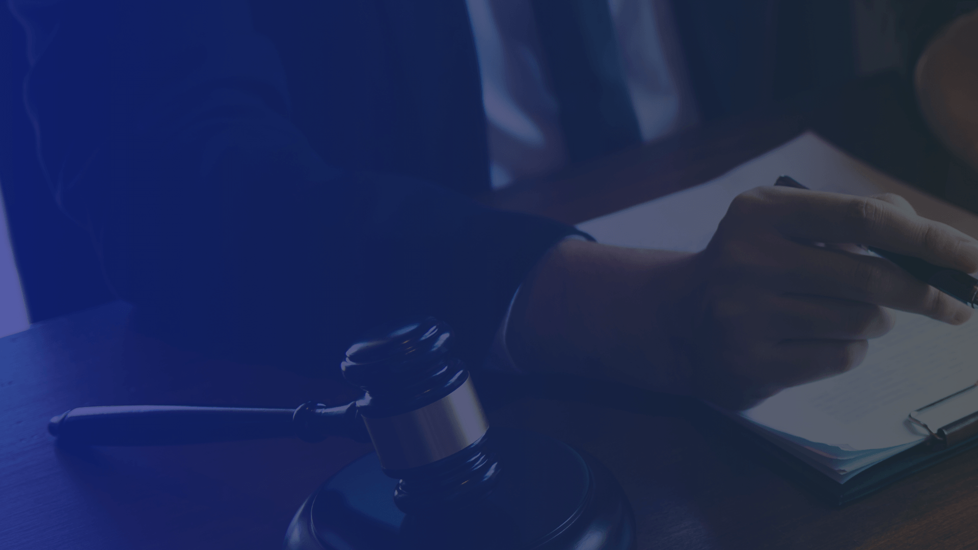 A gavel sits on a wooden desk next to a person's hand writing on a document.