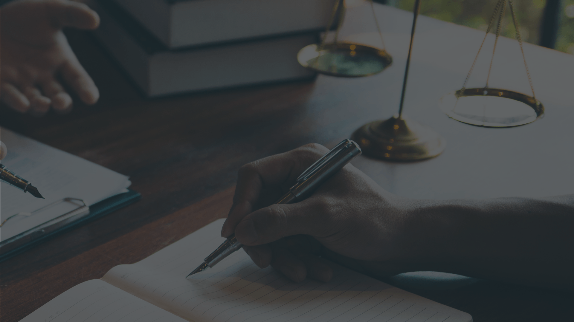 A hand writes in a notebook on a desk next to law books and a set of scales, signifying a professional legal setting.