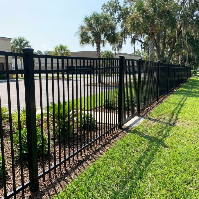 Black metal fence along a grassy roadside with trees and a parking area behind it