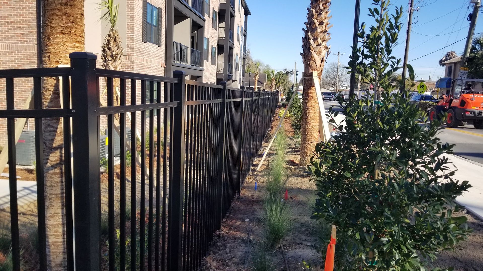 Black metal fence beside apartment buildings, palm trees, and landscaped walkway on a sunny day