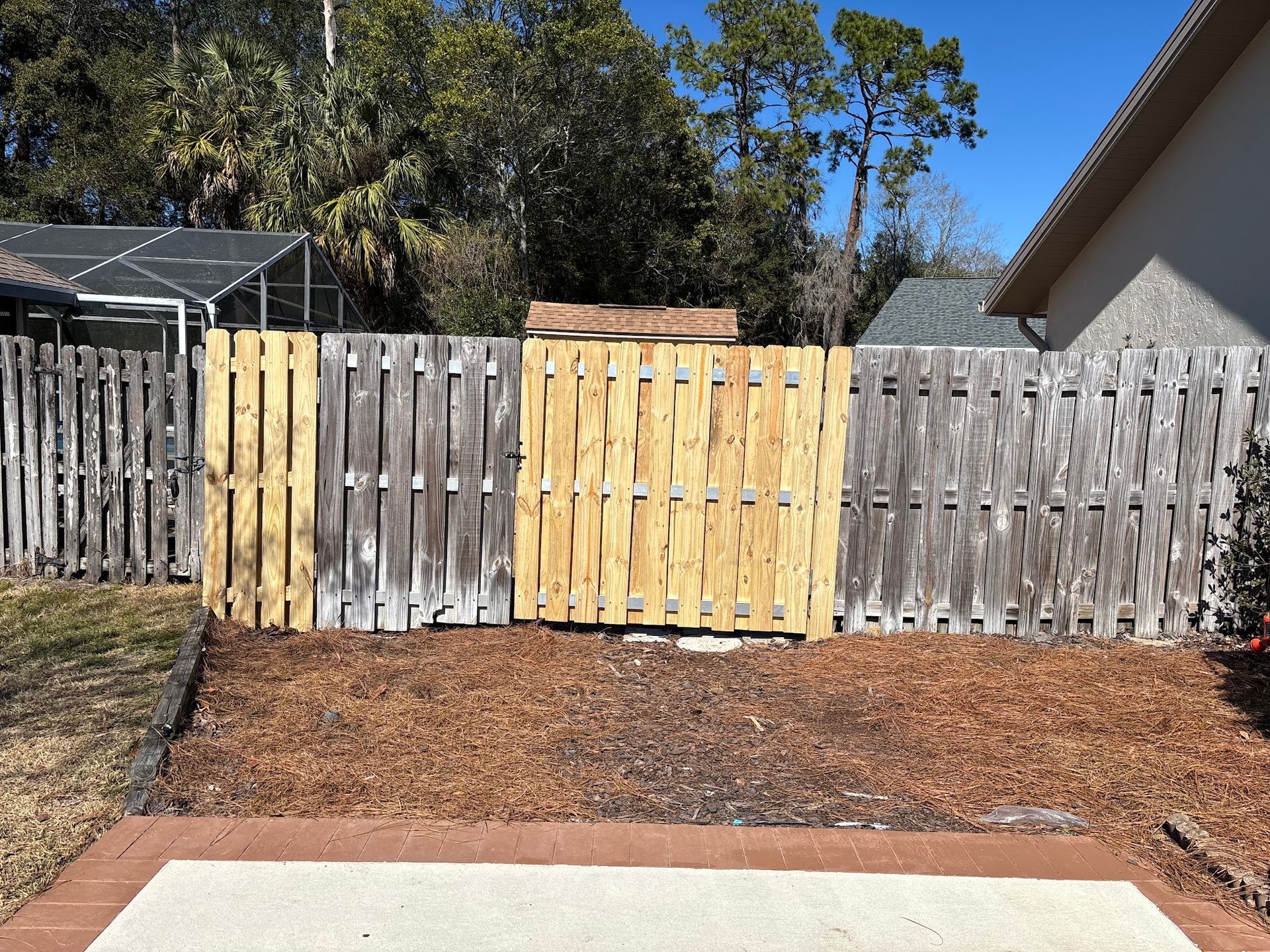Weathered wooden fence with a new pale-yellow gate and mulched garden bed in a backyard.